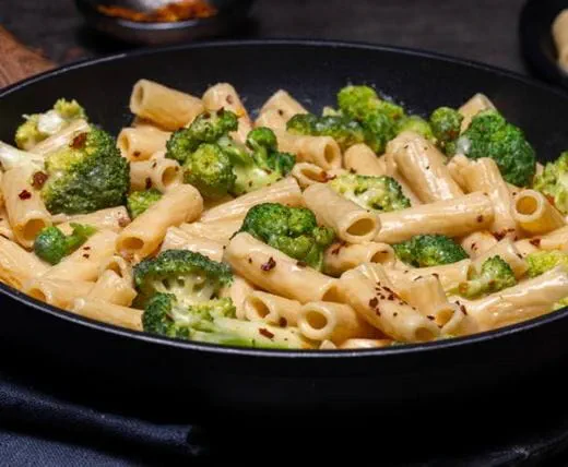 A skillet filled with rigatoni pasta mixed with broccoli florets and a creamy dairy free cheddar-style sauce, topped with red pepper flakes.