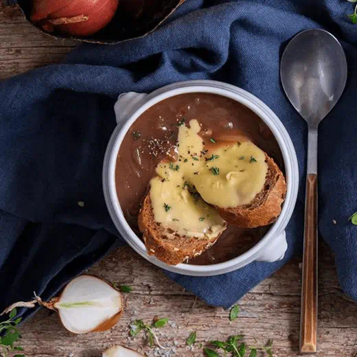 French onion soup topped with toasted bread and melted dairy free cheese and herbs, set on a rustic wooden table.