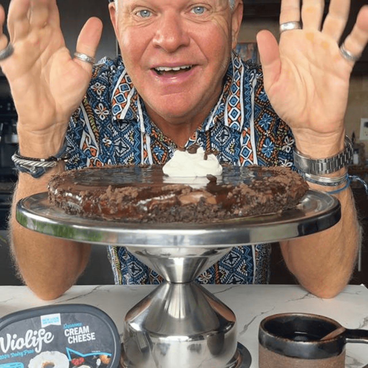 No‑bake espresso martini cheesecake on a metal cake stand, topped with whipped cream, chef standing in the background with hands raised.