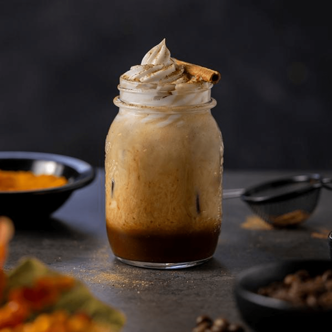 Pumpkin spice coffee in a mason jar topped with dairy free cream and a cinnamon stick, surrounded by coffee beans, pumpkin purée, and fall spices.