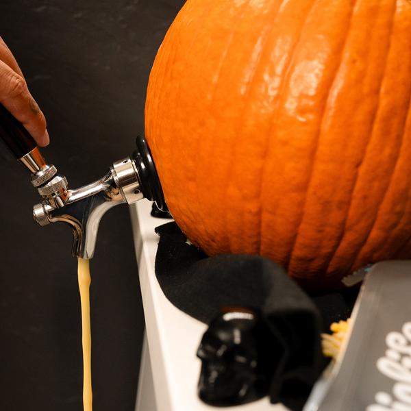 Large pumpkin fitted with a metal tap dispensing warm dairy free queso onto a surface decorated with black Halloween accents and snacks.