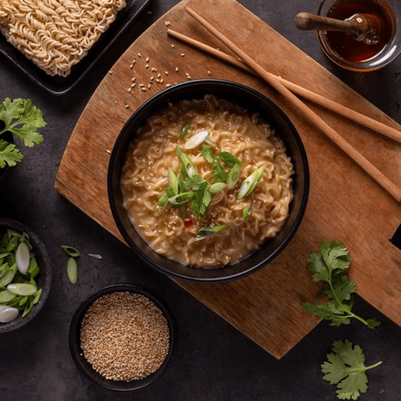 Bowl of spicy noodles topped with sliced green onions and cilantro, surrounded by uncooked ramen, sesame seeds and fresh herbs.