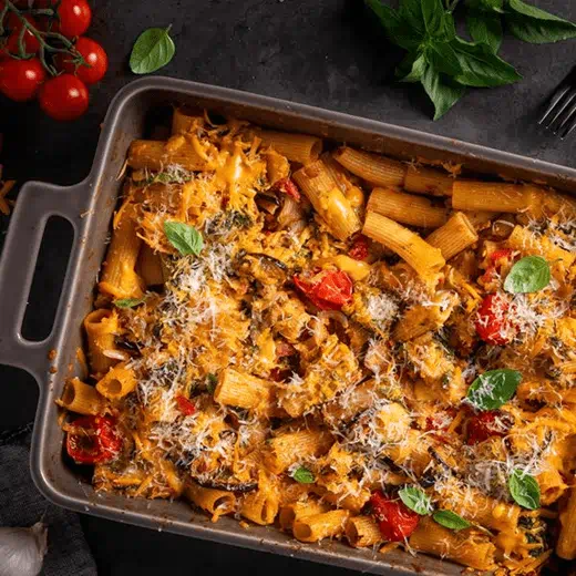 Baked eggplant pasta in a rectangular dish, featuring rigatoni mixed with roasted eggplant, cherry tomatoes, basil and melted dairy free cheese.