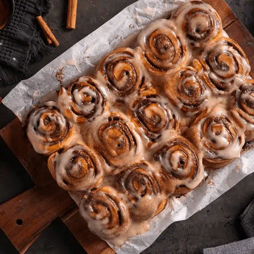 Freshly baked homemade cinnamon rolls covered in creamy icing, arranged on parchment paper over a wooden board with cinnamon sticks.