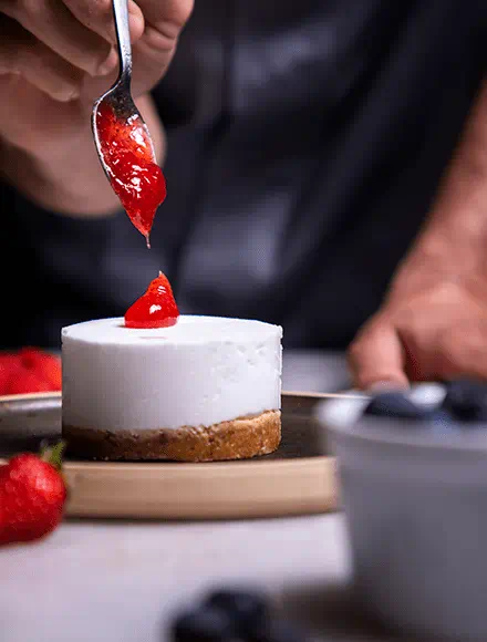 A hand holding a spoon adds a dollop of bright red strawberry sauce on top of a small round cheesecake with a crumb crust. Fresh strawberries and blueberries are placed around the plate.