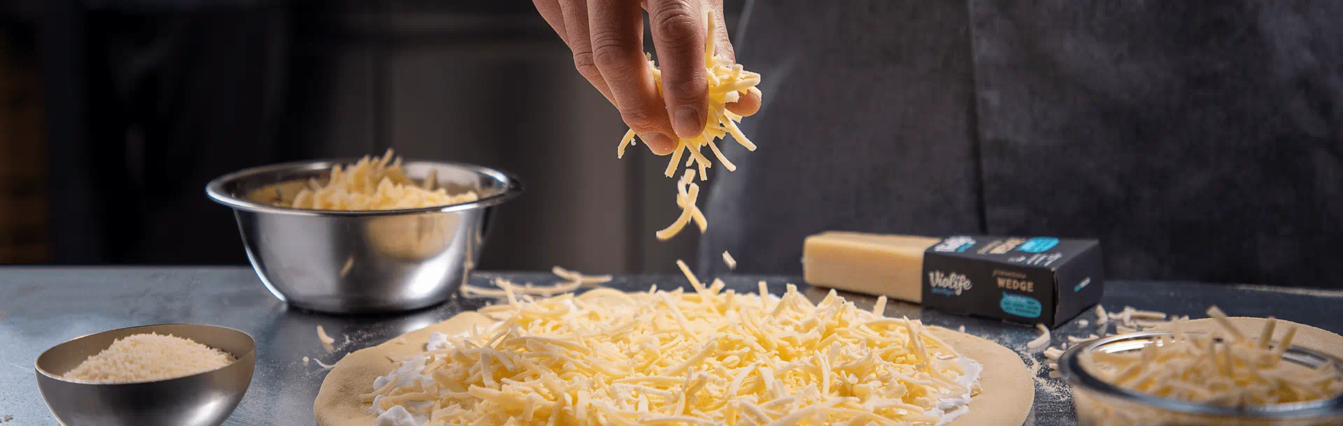 Hand sprinkling shredded Violife dairy-free cheese onto a pizza base on a countertop. Several bowls of grated Violife, a dairy free block of cheese and its packaging are visible around the pizza.