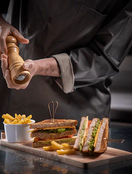 Hands seasoning a serving of French fries with a pepper grinder next to a wooden board holding a club sandwich filled with lettuce and tomato, accompanied by more fries.