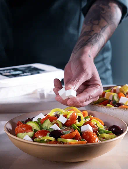 Hand adding small cubes of Violife Just Like Feta to a bowl of Greek salad with tomatoes, cucumbers, bell peppers and olives.