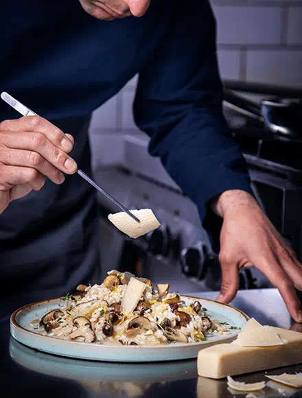 Hands plating a creamy pasta topped with mushrooms and garnished with Violife Just Like Parmesan on a round ceramic plate. 
