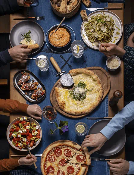 Groupe de personnes autour d’une table dégustant pizza, bruschetta, salade grecque, pâtes et dips préparés avec les produits Violife 100% vegan