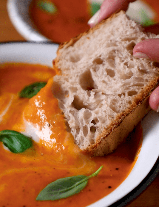 Main tenant une tranche de pain à tremper dans une soupe à la tomate réalisée avec Violife crémeux, l'alternative 100% végétale au fromage