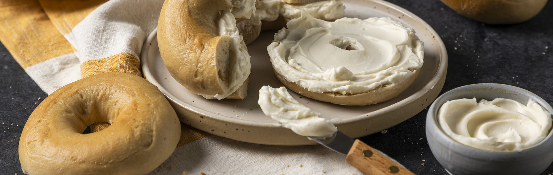 Un bagel coupé en deux et tartiné de Violife Supreme Crémeux, entouré de bagels entiers, d’un couteau à tartiner et d’un petit bol de tartinable Violife.