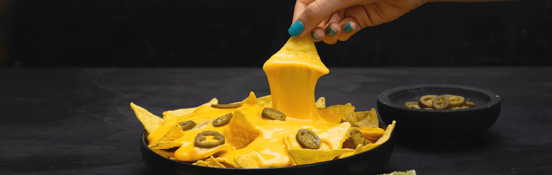 Main trempant une tortilla dans une sauce chaude de Violife, l'alternative végétale au fromage, sans produits laitiers, avec nachos et rondelles de jalapeño en dessous